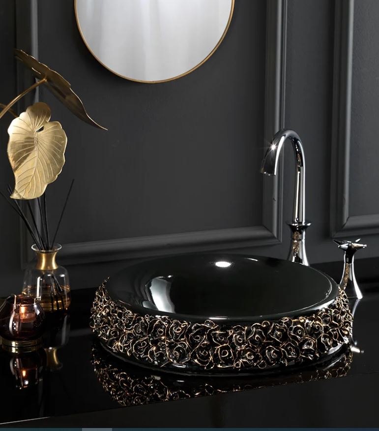 A glossy black round washbasin with gold floral patterns around the edge, accompanied by a silver faucet, set against a dark background with a golden round mirror and decorative items.