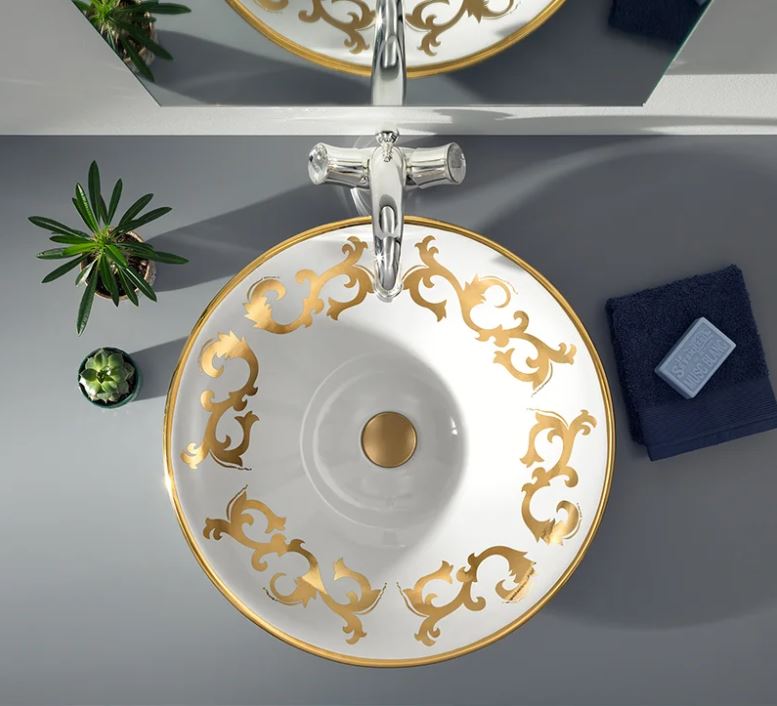 A white ceramic wash basin with gold spiral patterns, placed on a grey surface with a silver tap and green plants in the background.