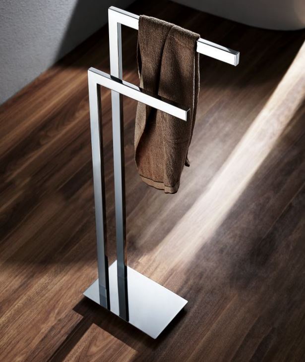 A silver stainless steel free standing towel bar with a towel hanging on it, placed on a wooden floor.