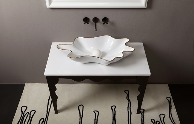 A white washbasin with gold detailing on the edges, placed on a white surface with a dark leg, against a grey wall, with a black and white rug on the floor.