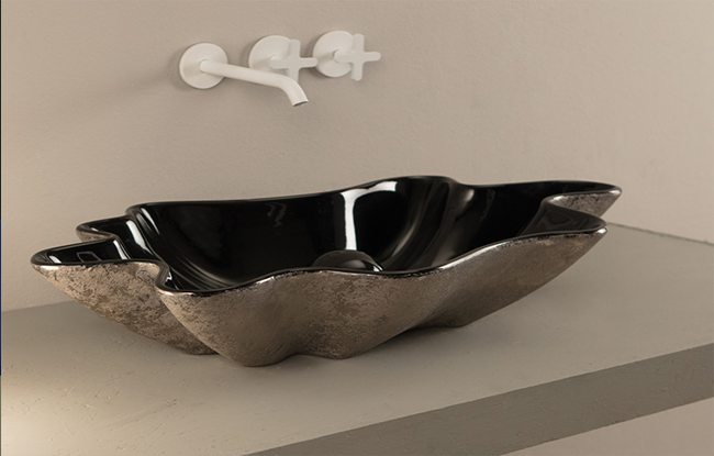 A black, uniquely shaped basin with a modern design, displayed in a bathroom setting with a wall-mounted tap above it.